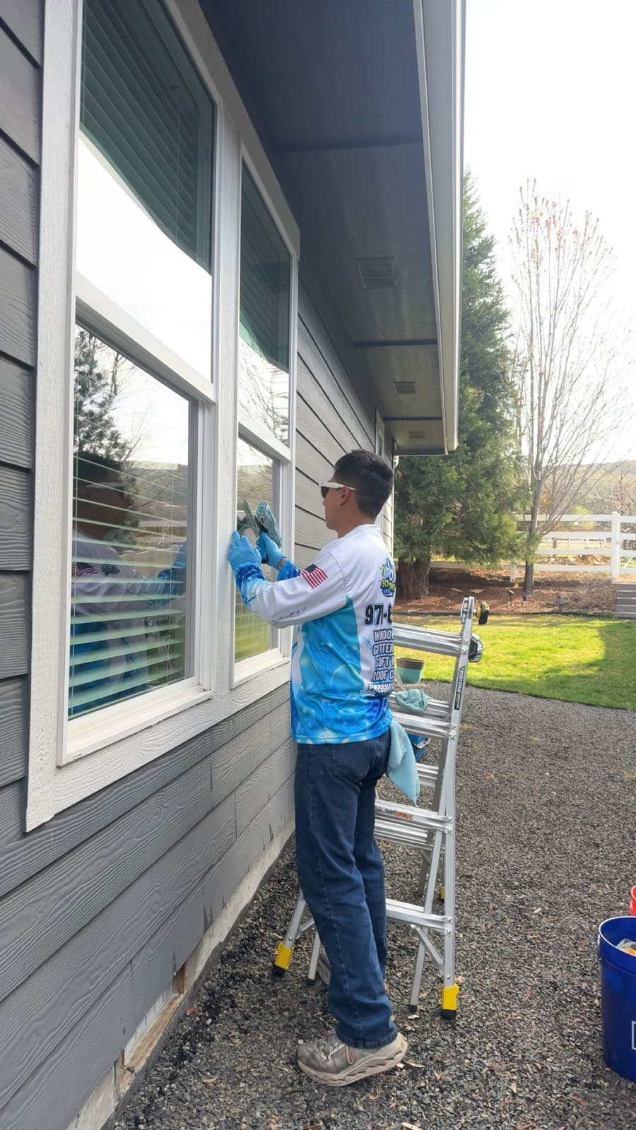 Technician cleaning windows from a ladder on a gray-siding home